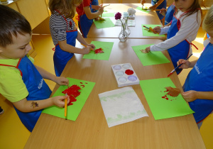 Dzieci stoją przy stoliku i malują liście czerwoną farbą